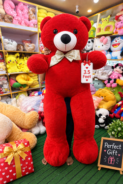A large red plush teddy bear with a white bow tie stands on a green floor inside a toy store, surrounded by shelves of other stuffed animals.